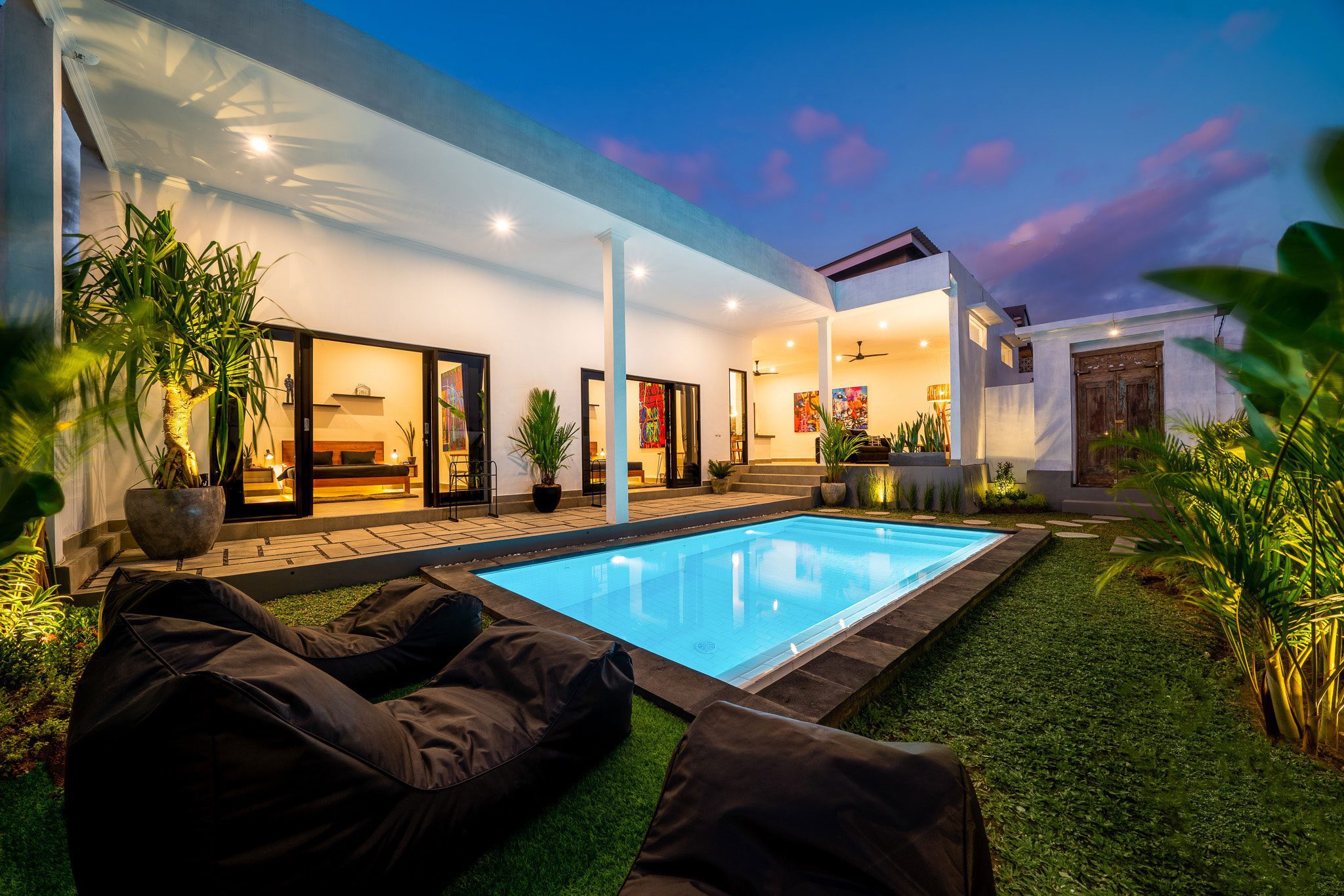 Outdoor view of a modern house with a lit rectangular pool, surrounded by greenery and outdoor seating, taken during twilight. Glass doors lead to the interior, and the sky has a mixture of blue and pink hues.